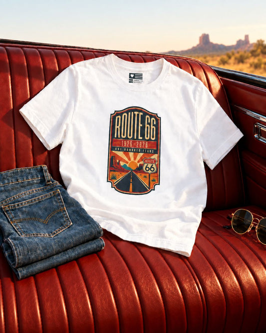 White Route 66 Centennial tee on a vintage red leather car seat beside denim and sunglasses, with desert scenery behind.
